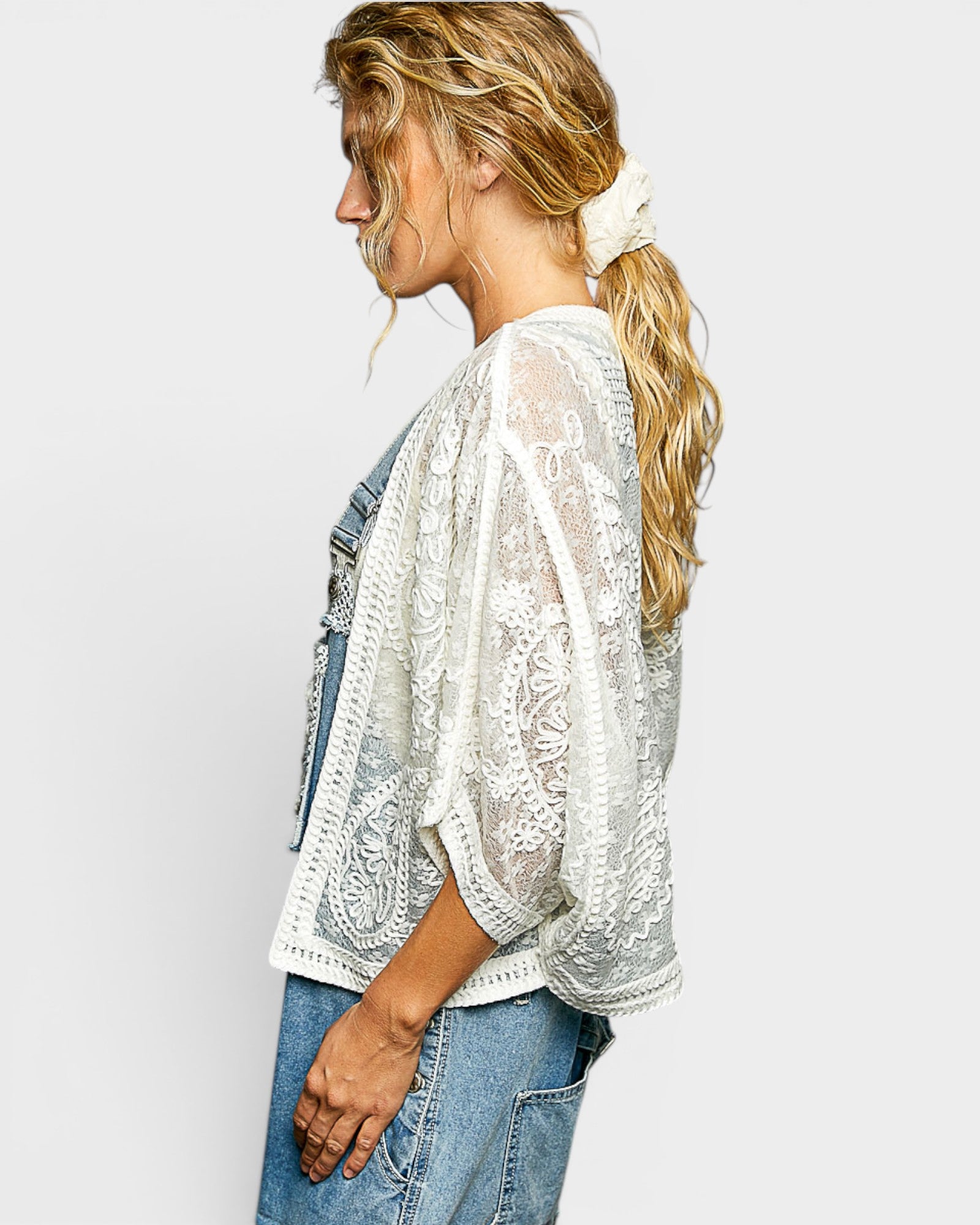 Woman wearing a white lace top and denim overalls on a plain background