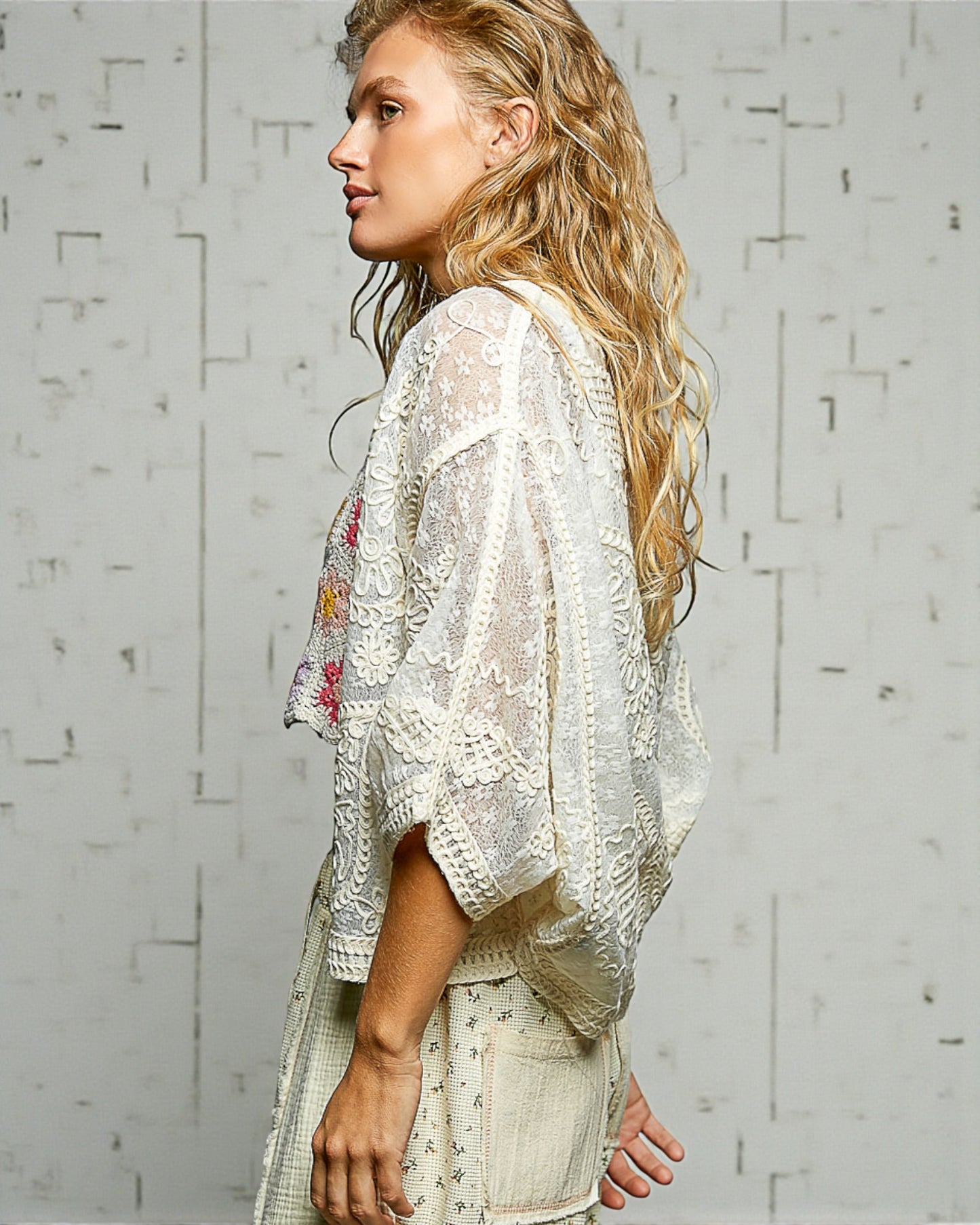 Woman wearing a white lace top with floral embroidery against a light gray brick wall.