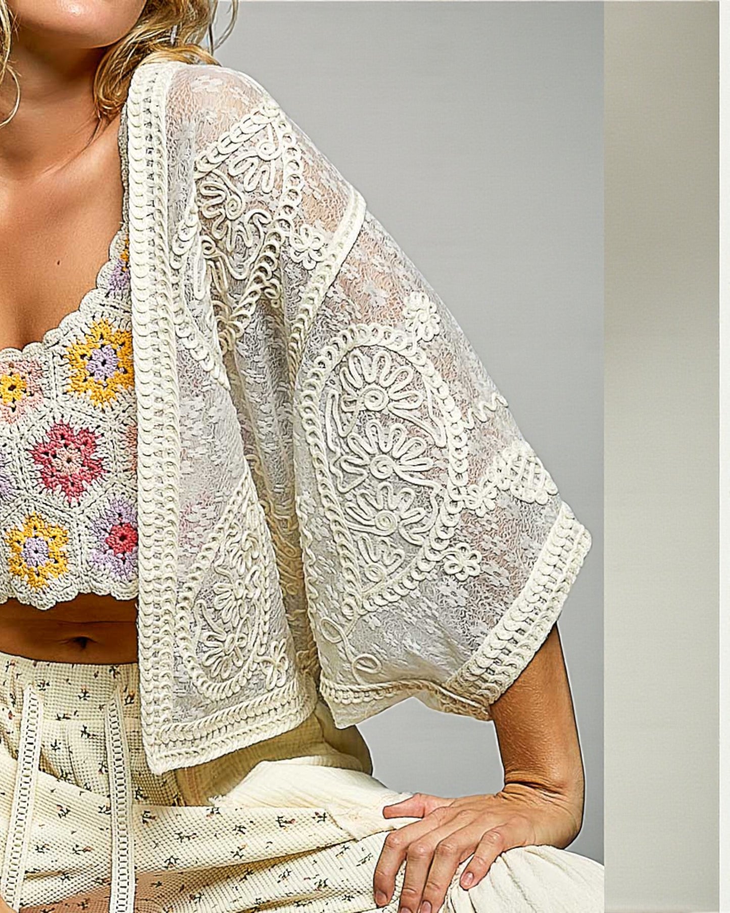 Woman wearing a white lace cover-up over a colorful floral top against a neutral background