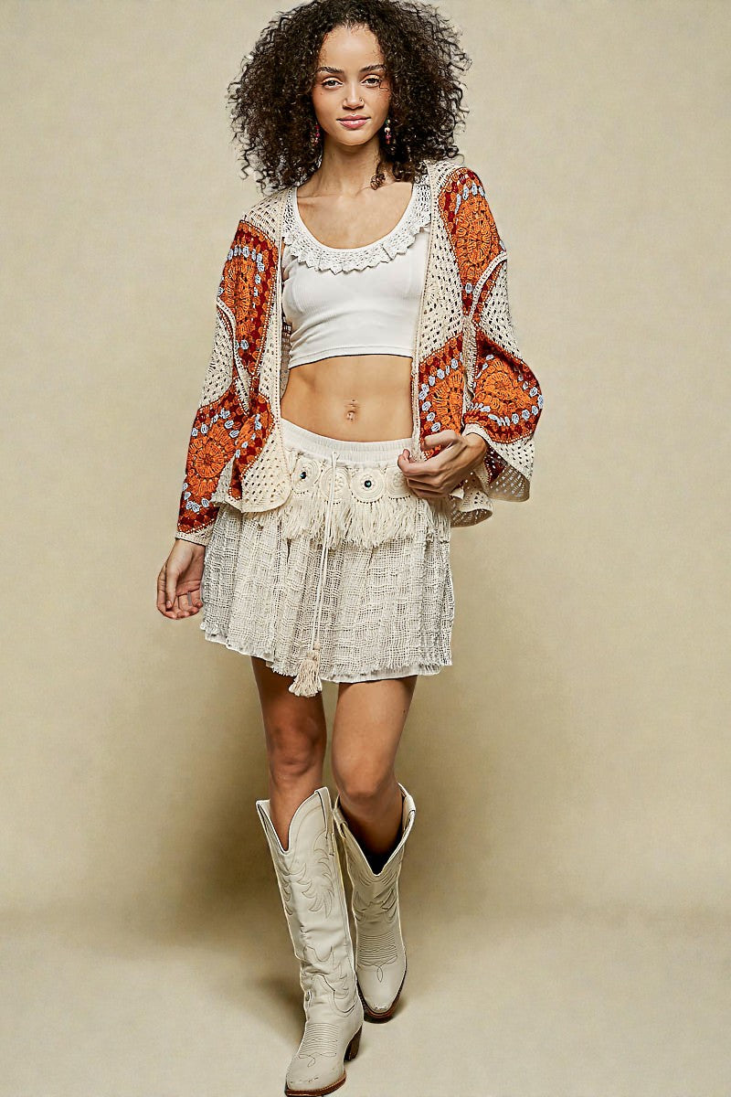 Woman wearing a patterned cardigan, white top, skirt, and cowboy boots against a beige background