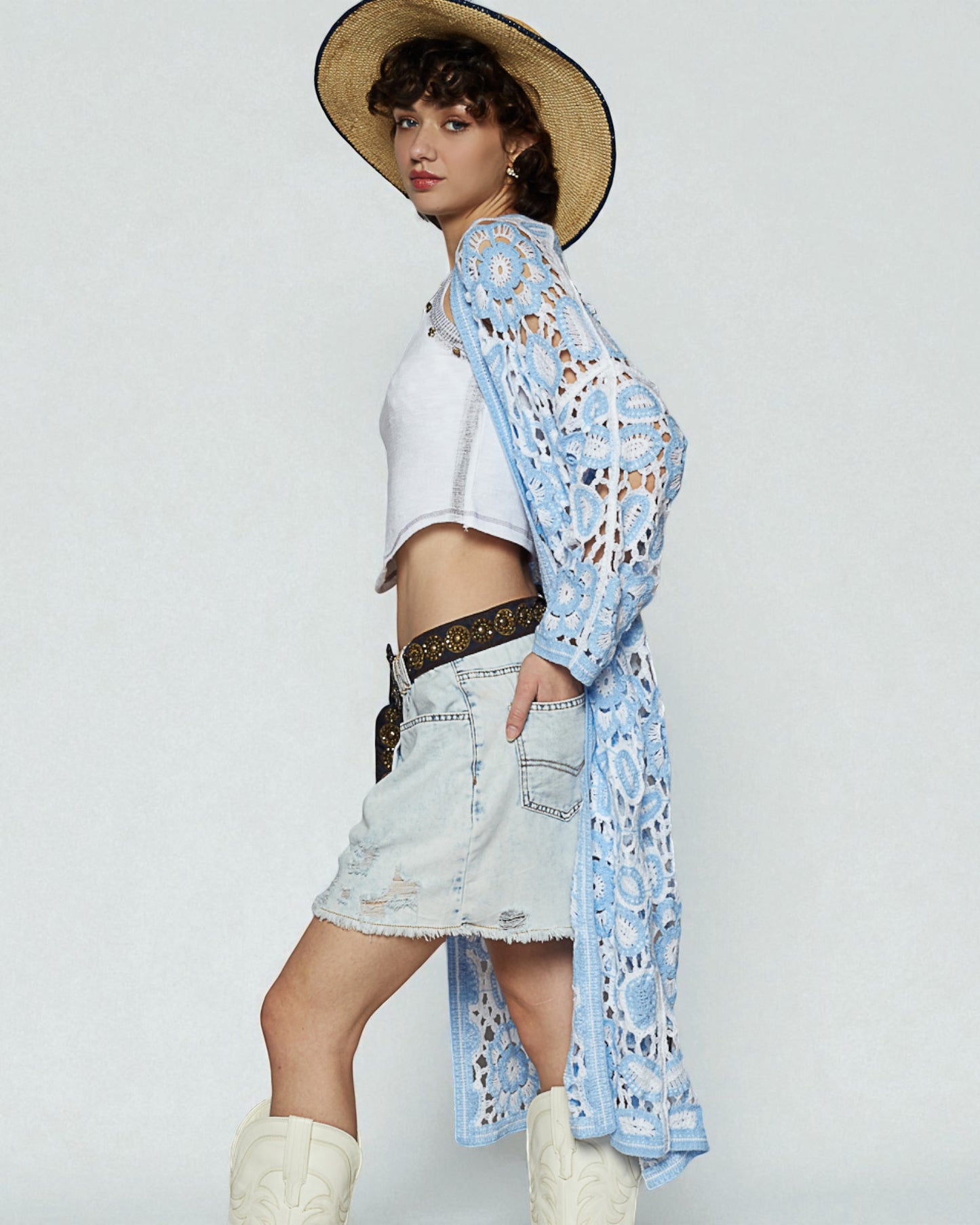 Woman wearing a white top, denim shorts, and a blue lace cover-up with a straw hat on a light gray background.