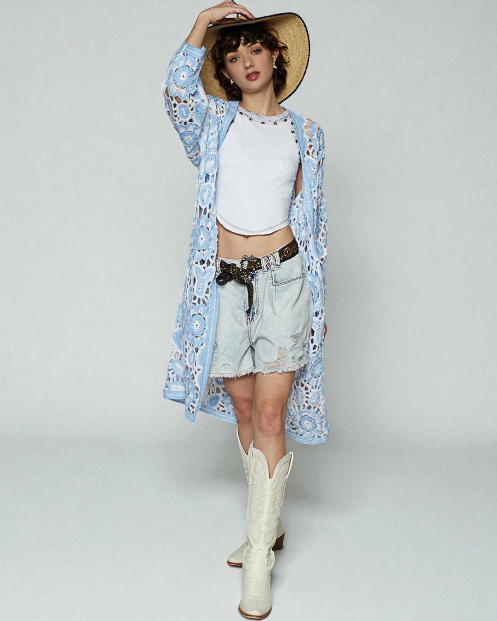 Woman wearing a blue floral kimono, white top, denim shorts, and white cowboy boots on a plain background