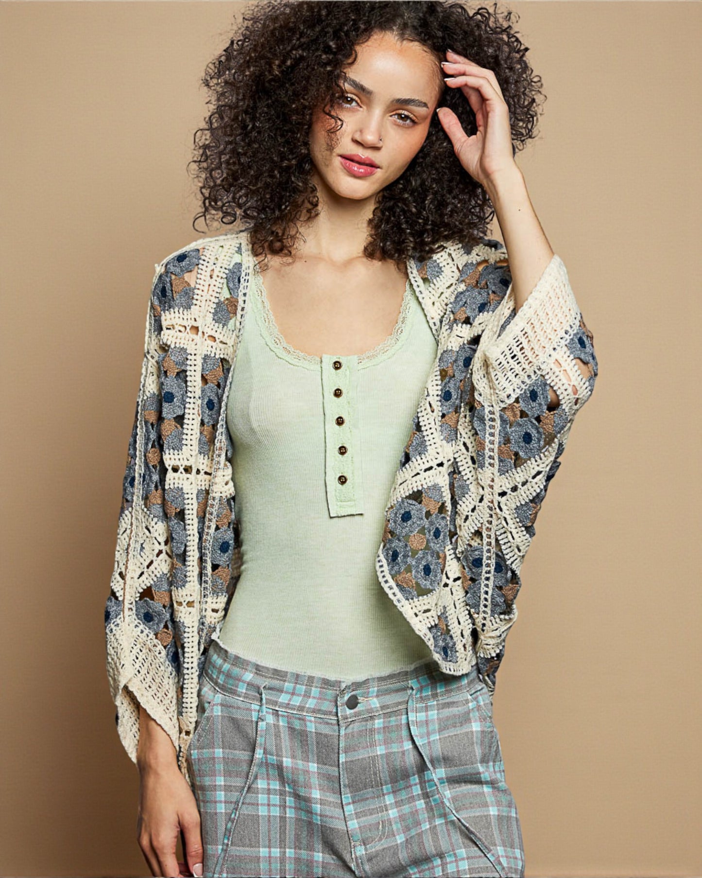 Woman wearing a floral crochet cardigan over a green top and plaid pants on a beige background