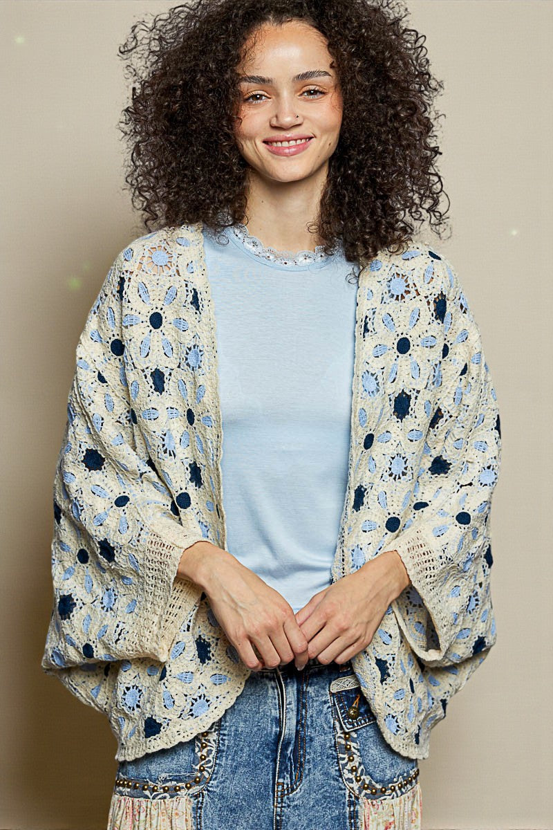 Woman wearing a floral crochet cardigan over a light blue shirt and jeans on a beige background