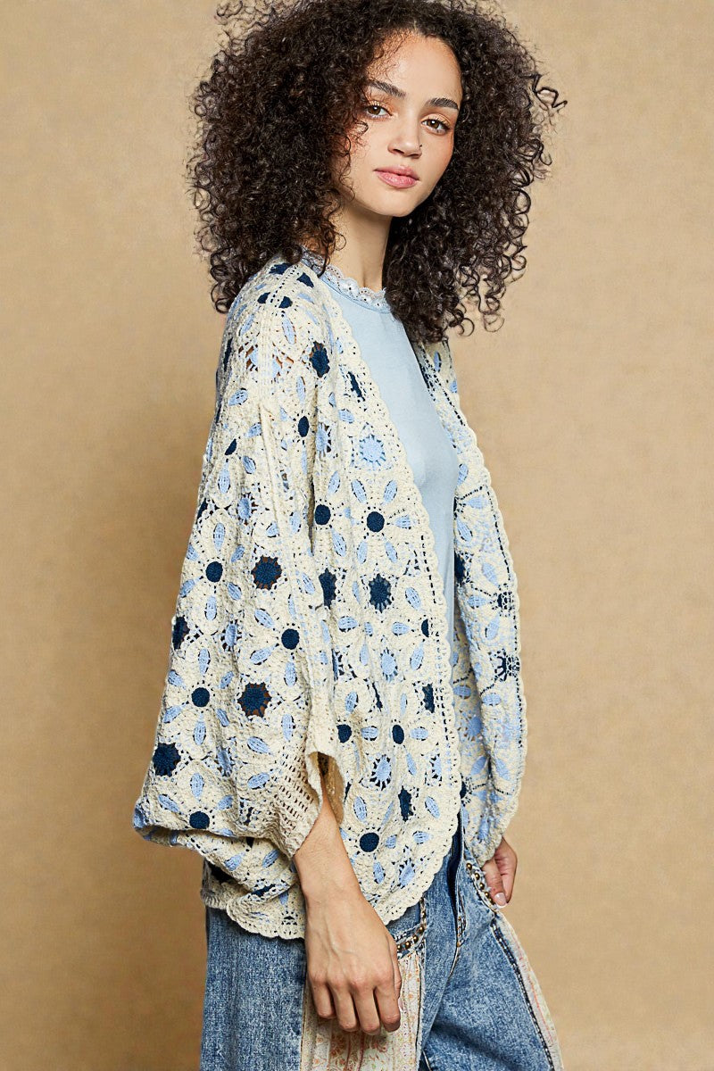 Woman wearing a floral crochet cardigan over a light blue shirt on a beige background
