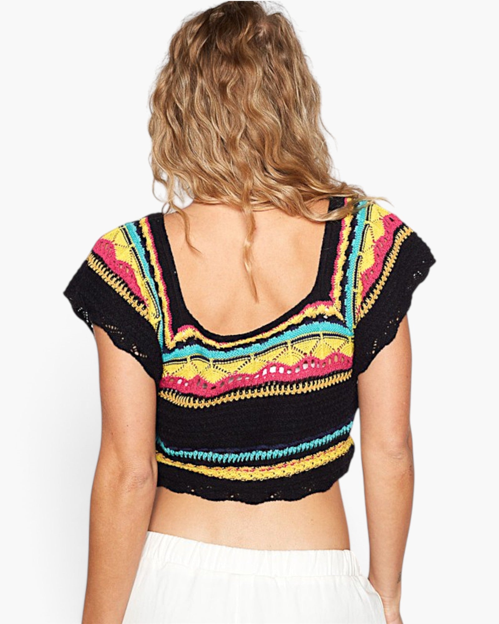 A woman viewed from the back wearing a black crocheted cropped top with colorful geometric stripes in pink, yellow, turquoise, and white, paired with white bottoms, against a plain white background.