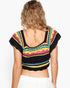 A woman viewed from the back wearing a black crocheted cropped top with colorful geometric stripes in pink, yellow, turquoise, and white, paired with white bottoms, against a plain white background.