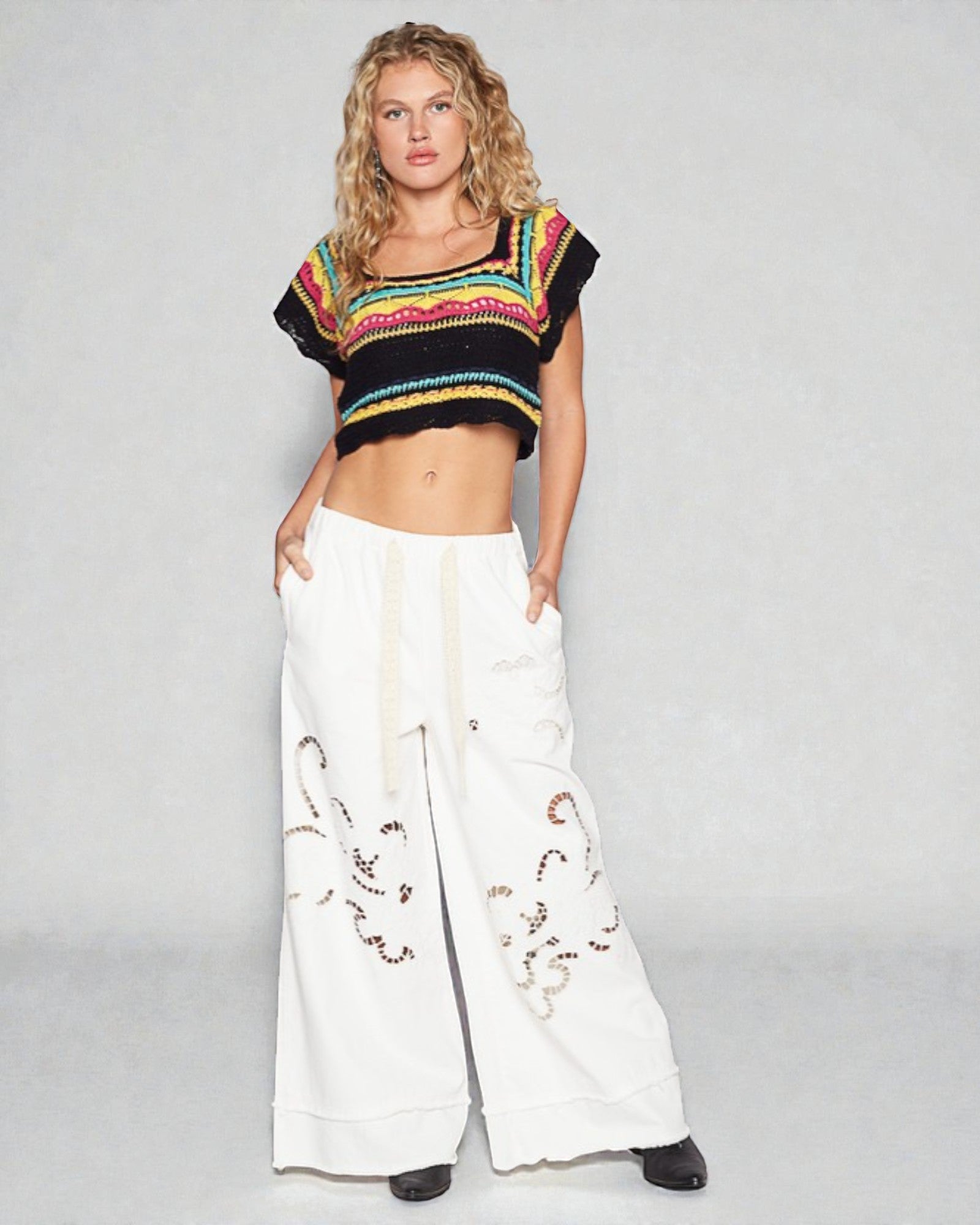 Woman wearing a colorful cropped top and white pants with embroidery on a gray background