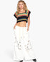 A woman modeling a black cropped knit top with colorful horizontal stripes in pink, yellow, green, and blue, paired with white wide-leg pants featuring embroidered details, against a plain white background.