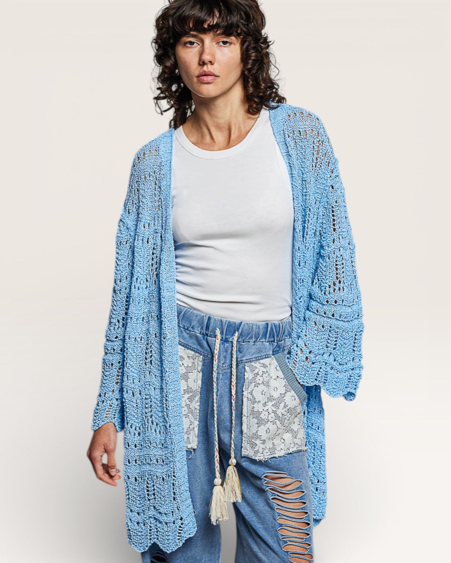 Woman wearing a light blue crochet cardigan over a white top and jeans with lace details.