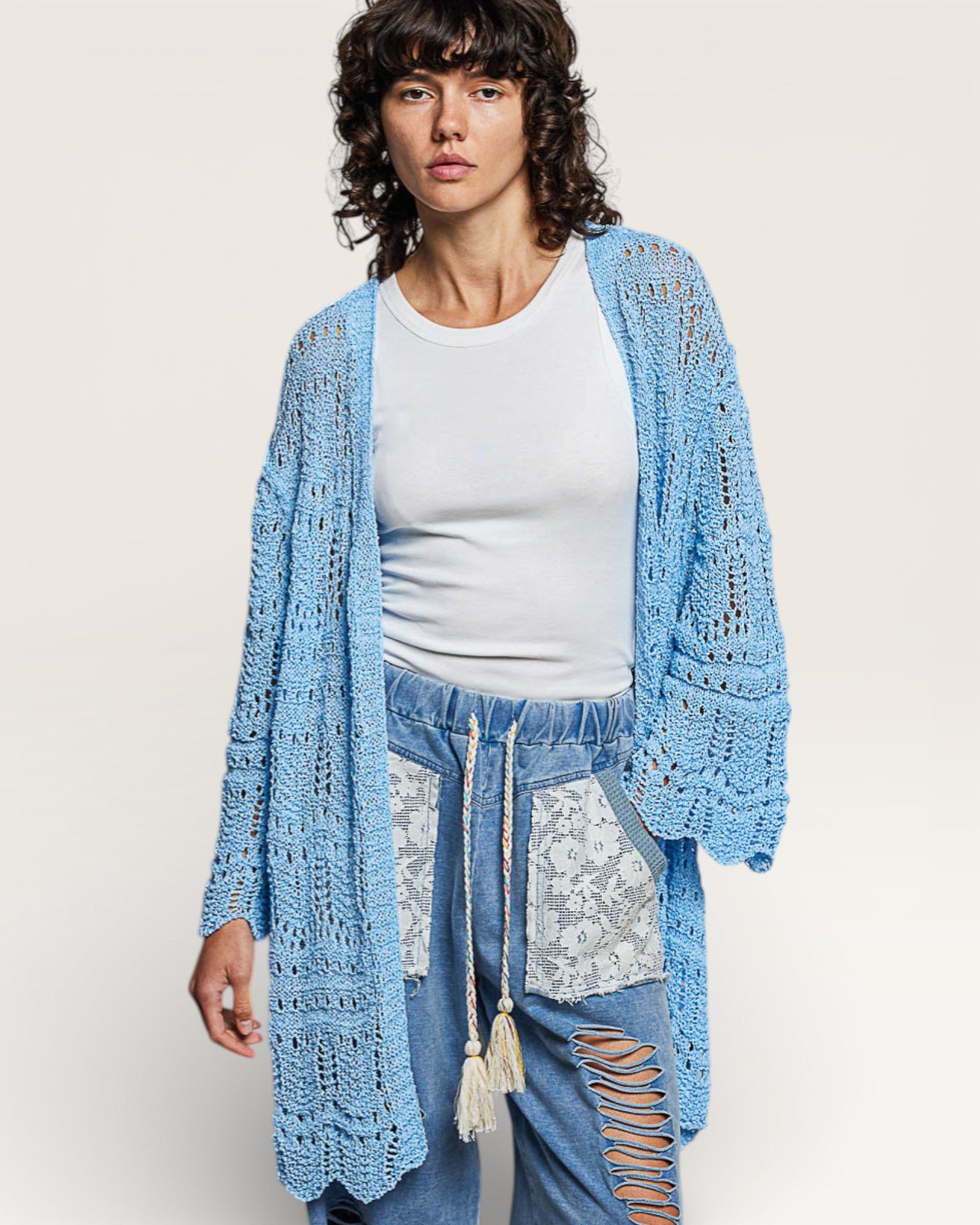 Woman wearing a light blue crochet cardigan over a white top and jeans with lace details.
