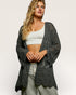 Woman wearing a dark gray crochet cardigan over a white top and light pants on a plain background