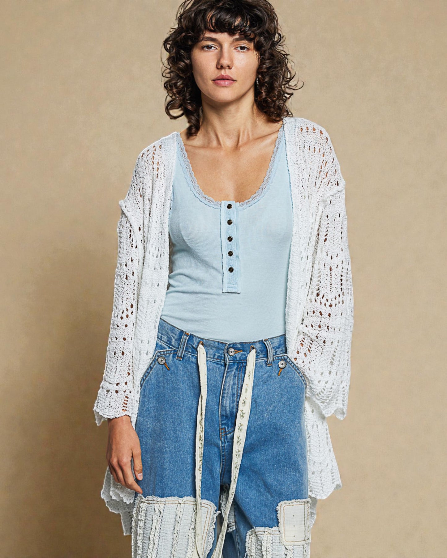 Woman wearing a light blue top and denim jeans with a white lace cardigan against a beige background
