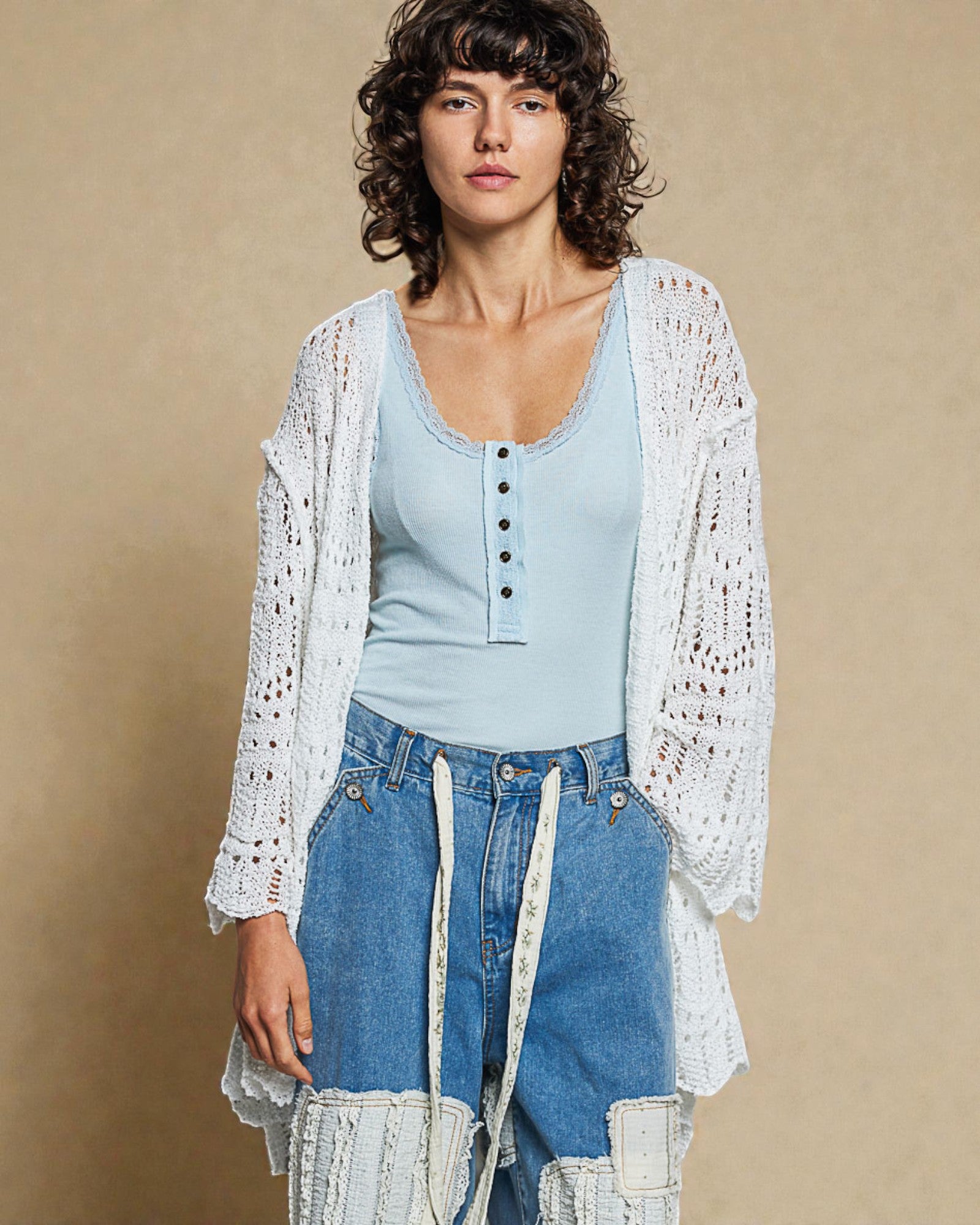 Woman wearing a light blue top and denim jeans with a white lace cardigan against a beige background