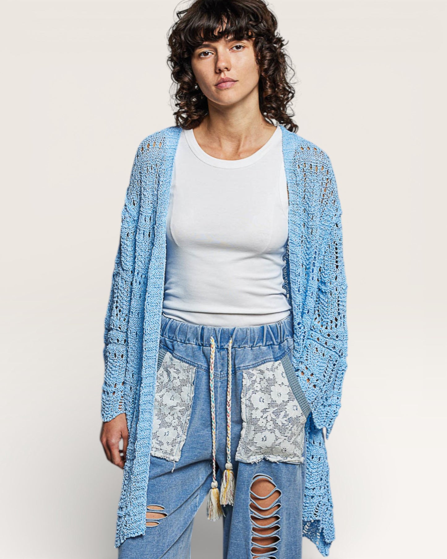 Woman wearing a light blue crochet cardigan over a white top and ripped jeans on a plain background