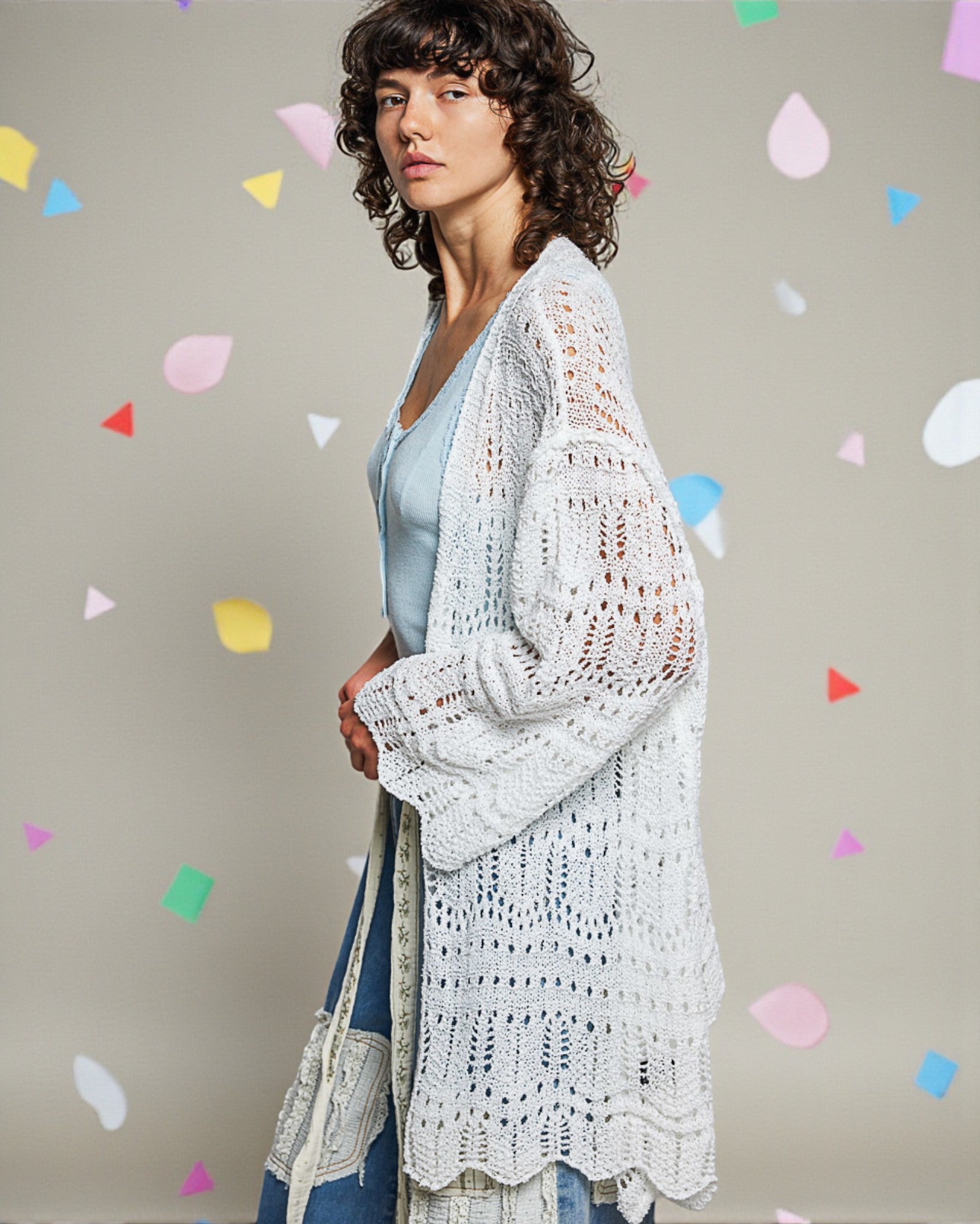 Woman wearing a white crochet cardigan over a blue top with colorful confetti shapes on a gray background
