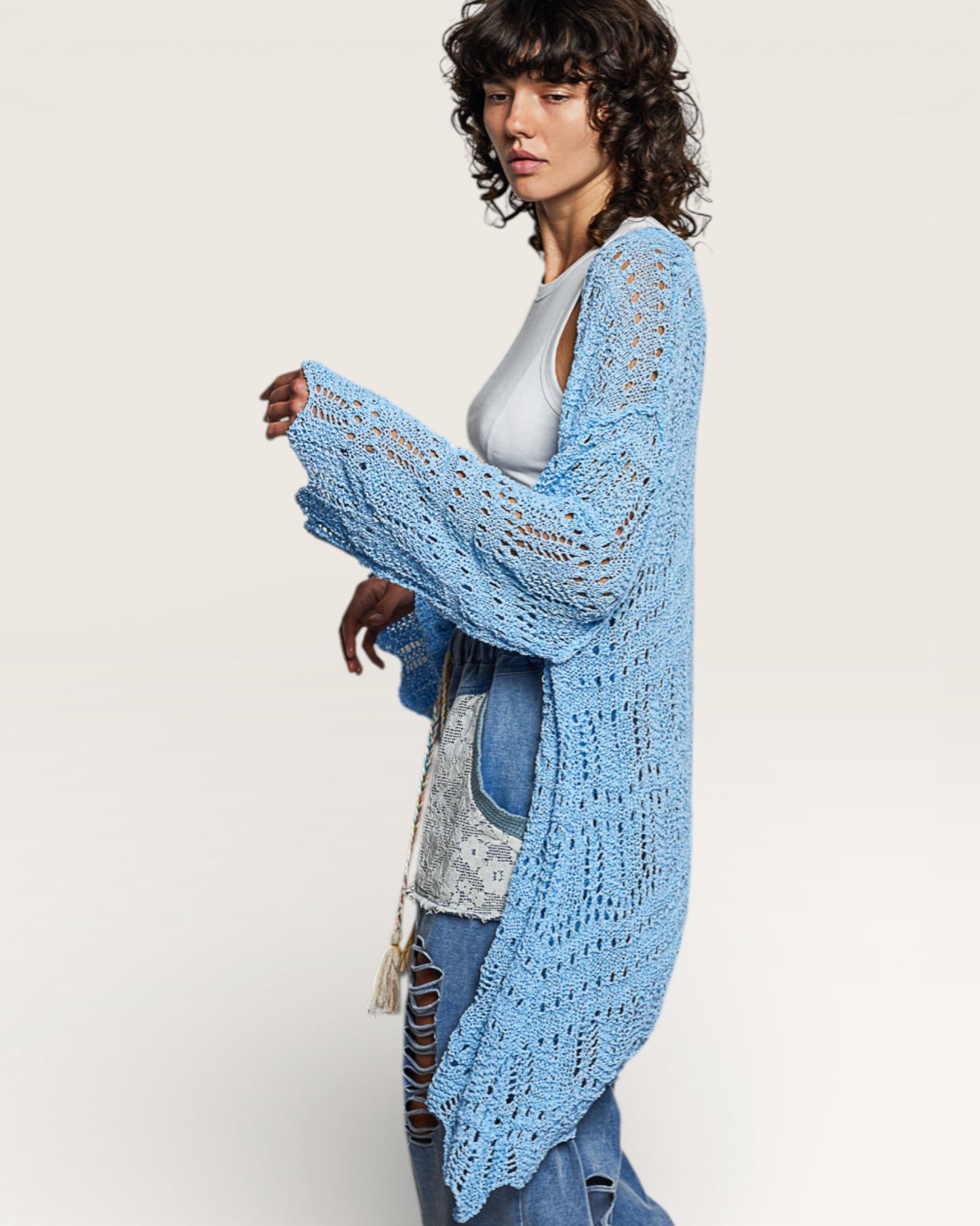 Woman wearing a light blue crochet cardigan over a white top and denim jeans on a plain background