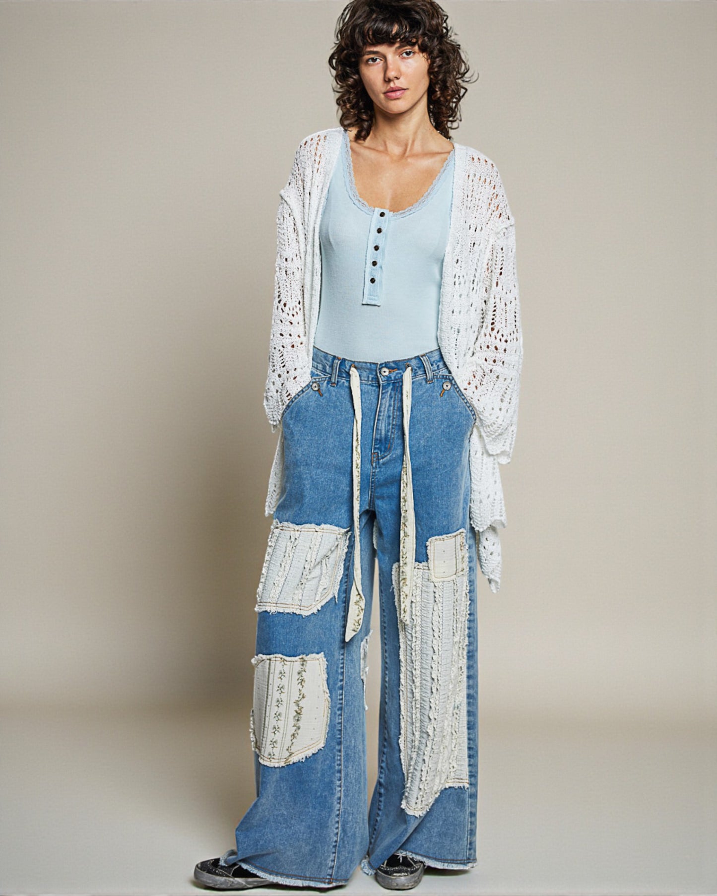 Woman wearing a light blue top, white lace cardigan, and patchwork jeans against a neutral background