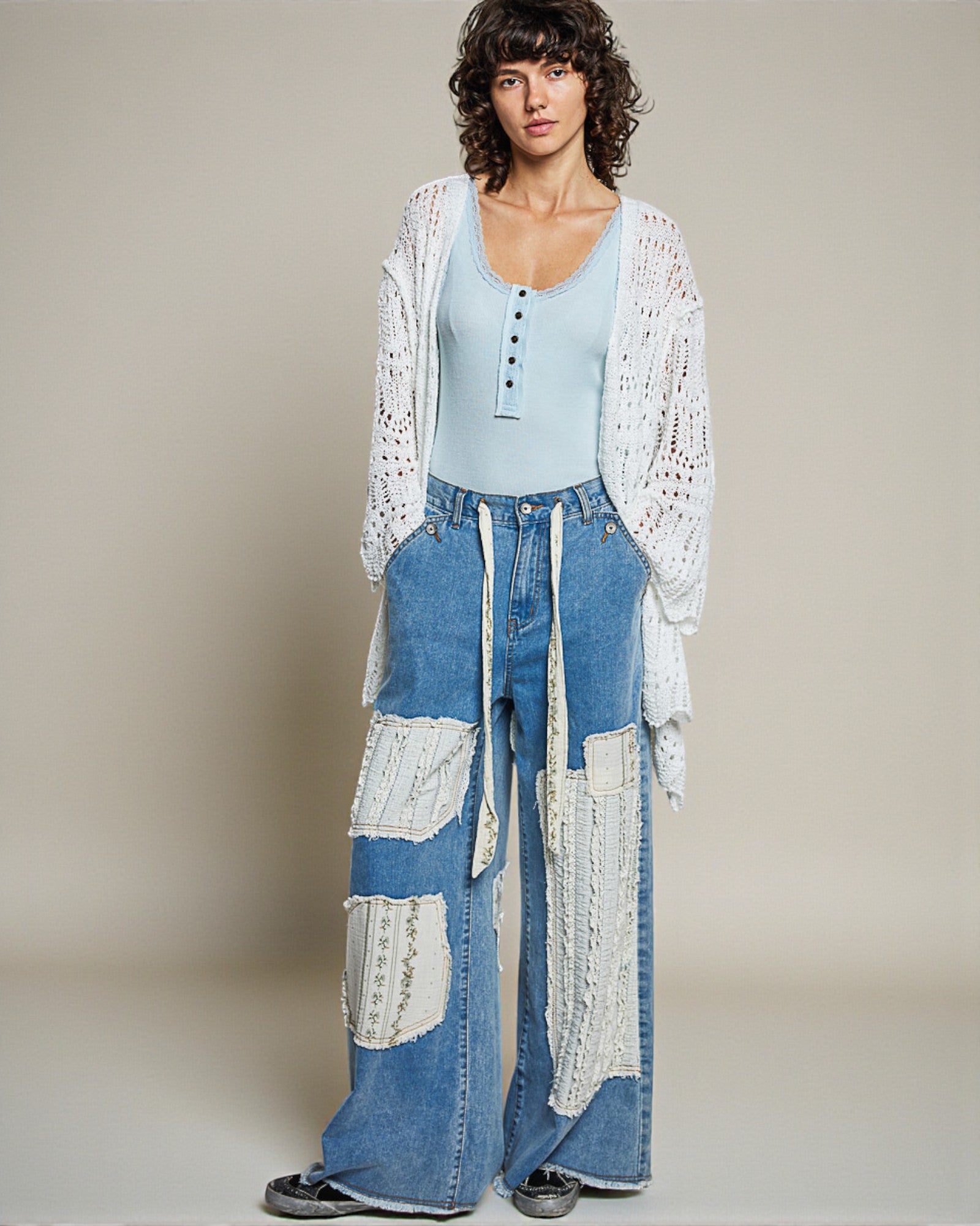 Woman wearing a light blue top, white lace cardigan, and patchwork jeans against a neutral background
