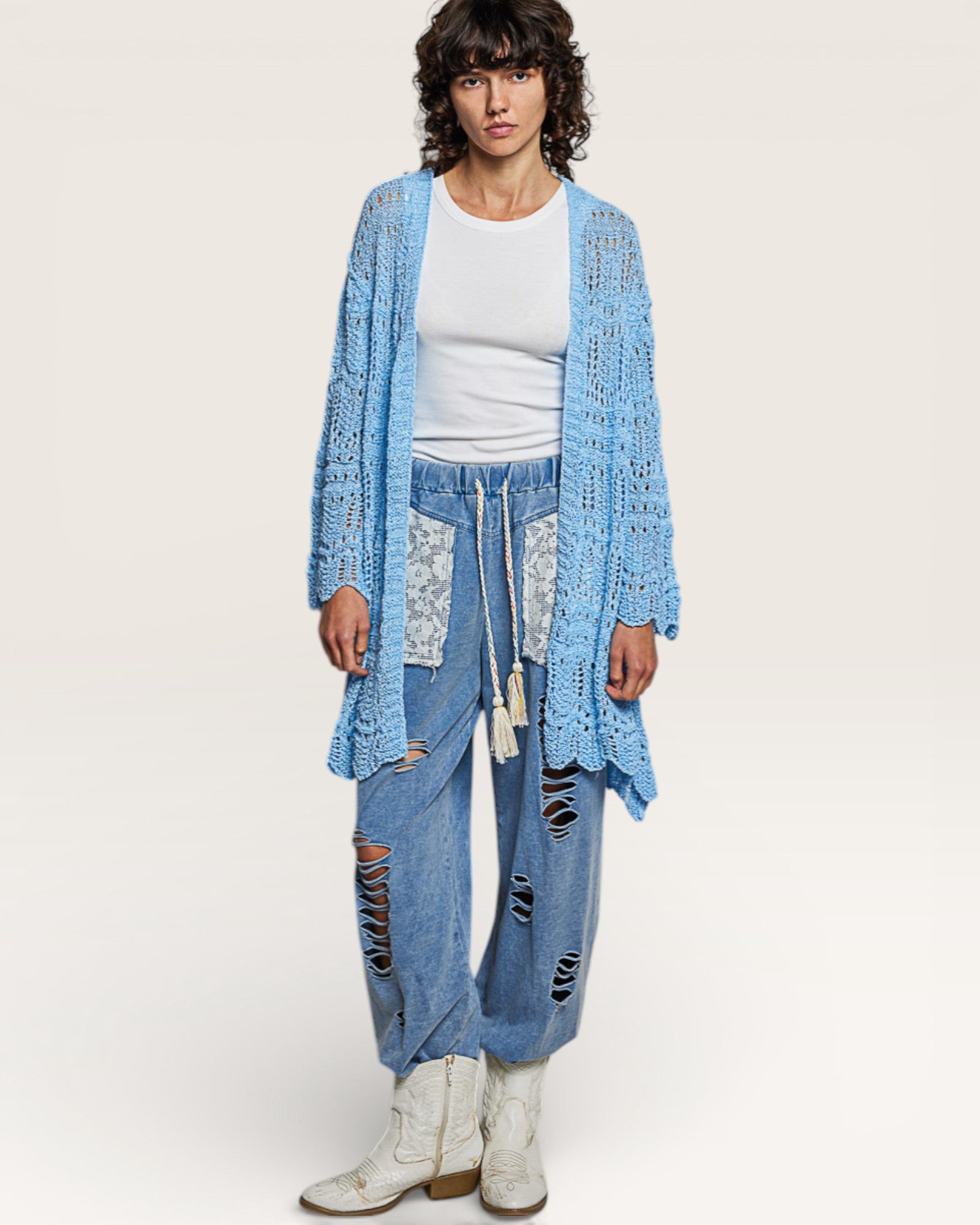 Person wearing a blue crochet cardigan, white top, and distressed denim pants on a white background