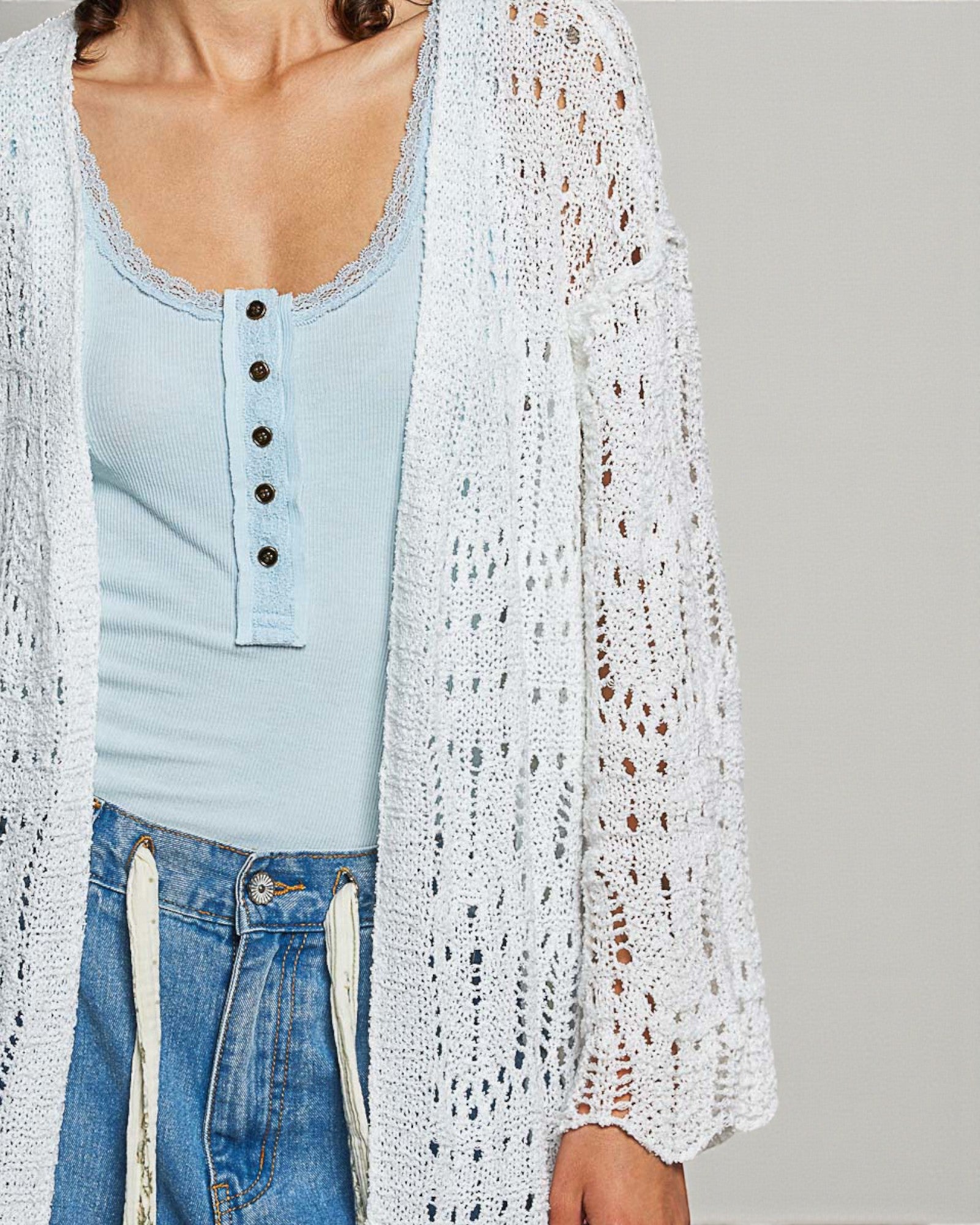 Person wearing a light blue button-up shirt, white crochet cardigan, and blue jeans on a plain background