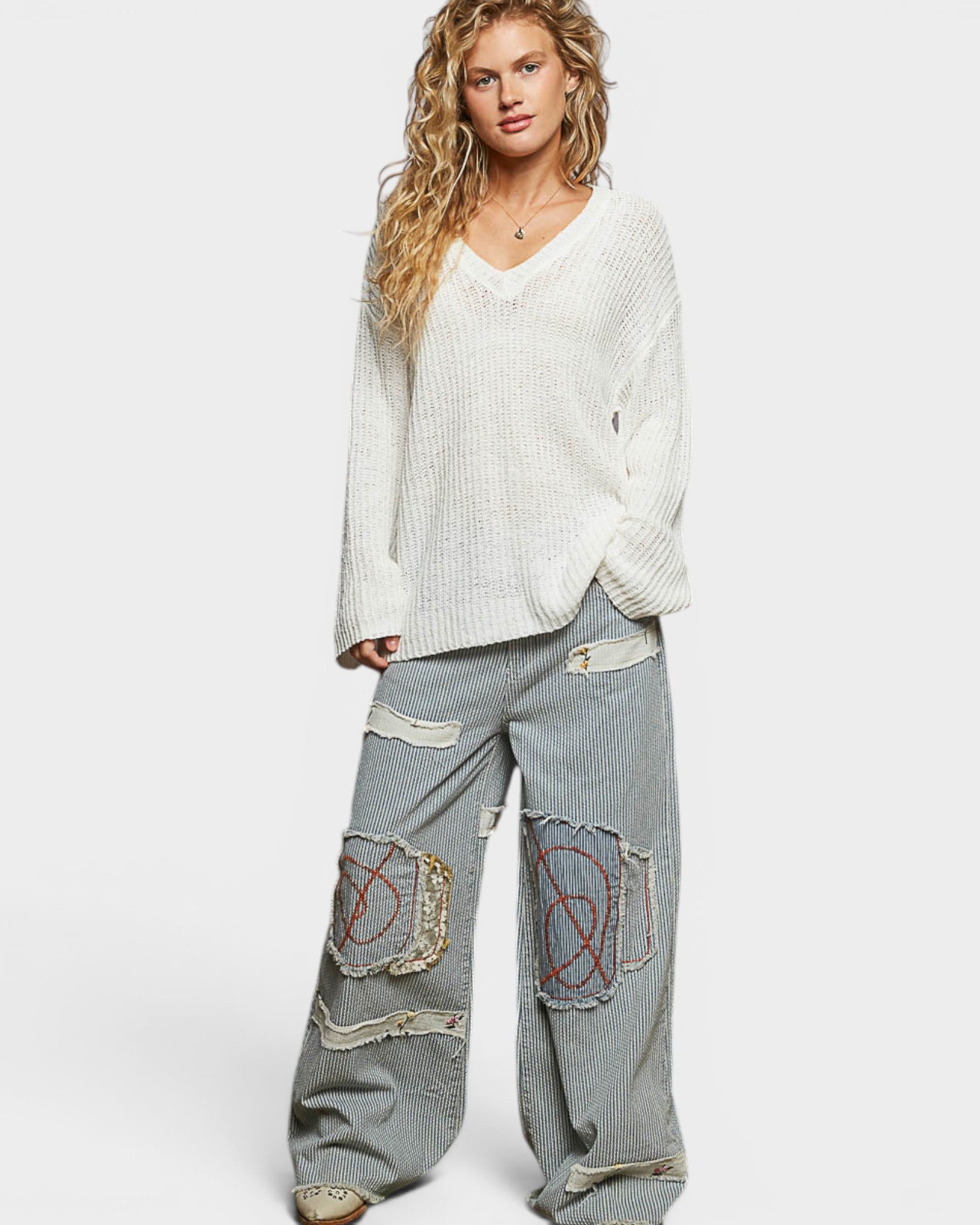 Woman wearing a white sweater and gray jeans with decorative patches on a white background
