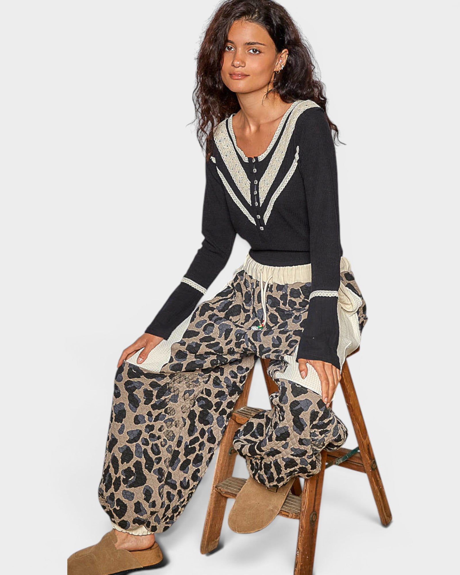Woman wearing a black sweater with white trim and leopard print pants, sitting on a wooden stool.