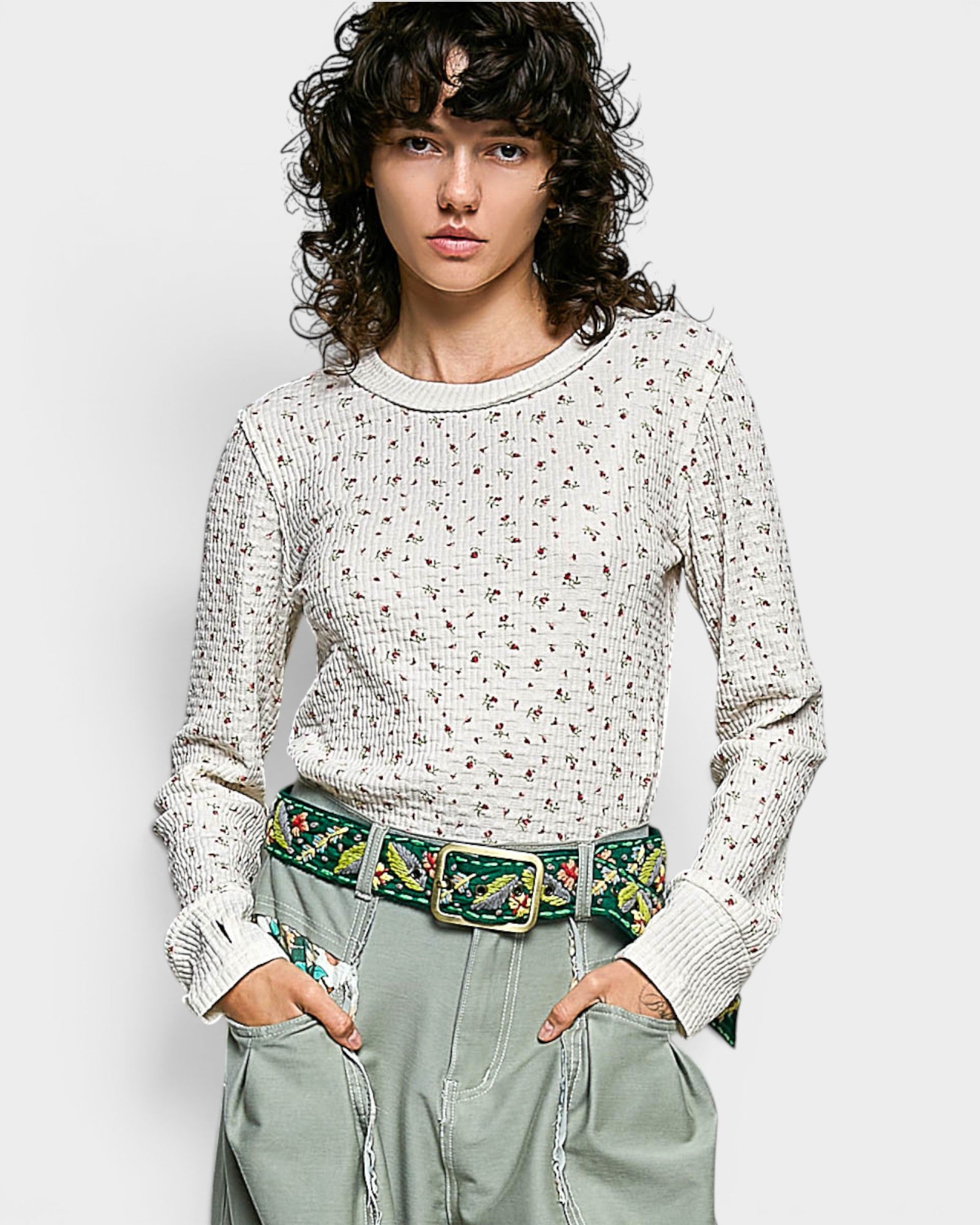 Person wearing a patterned sweater and green pants with a floral belt on a white background