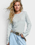 Woman wearing a light gray textured sweater and light blue jeans with a colorful belt on a white background