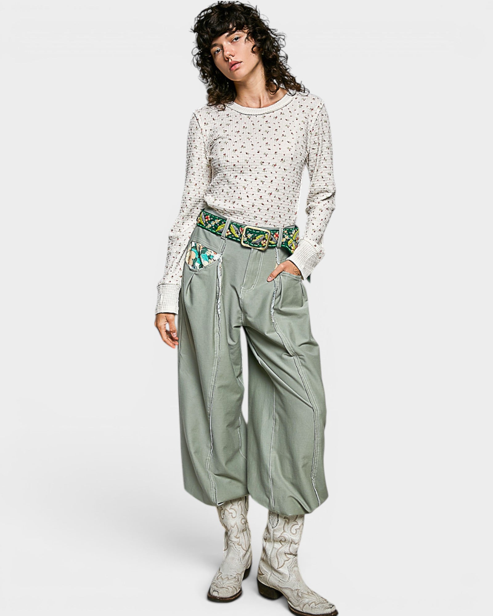 Woman wearing a patterned top and green pants with embroidery, standing against a white background