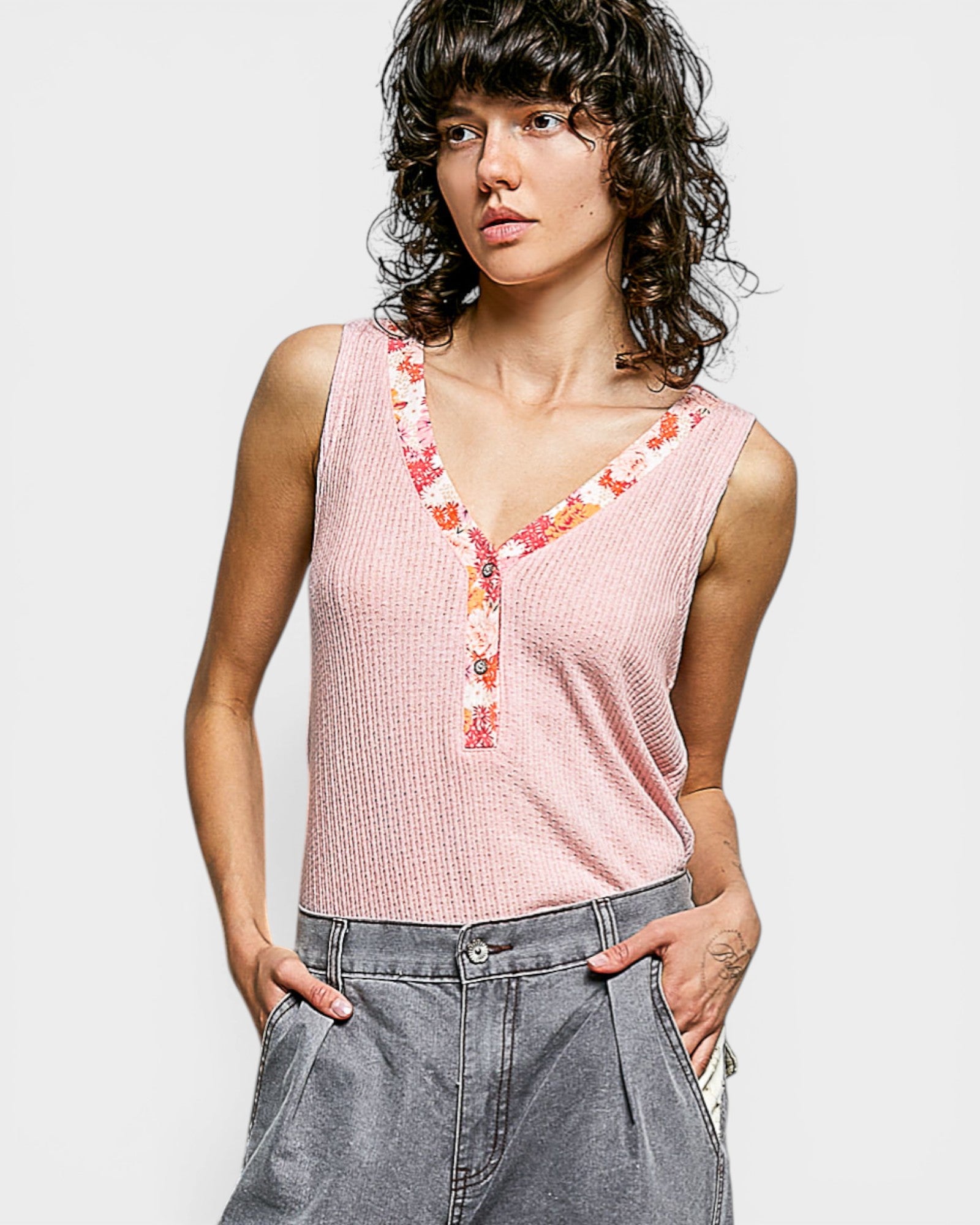 Woman wearing a pink sleeveless top with floral details and gray pants on a white background