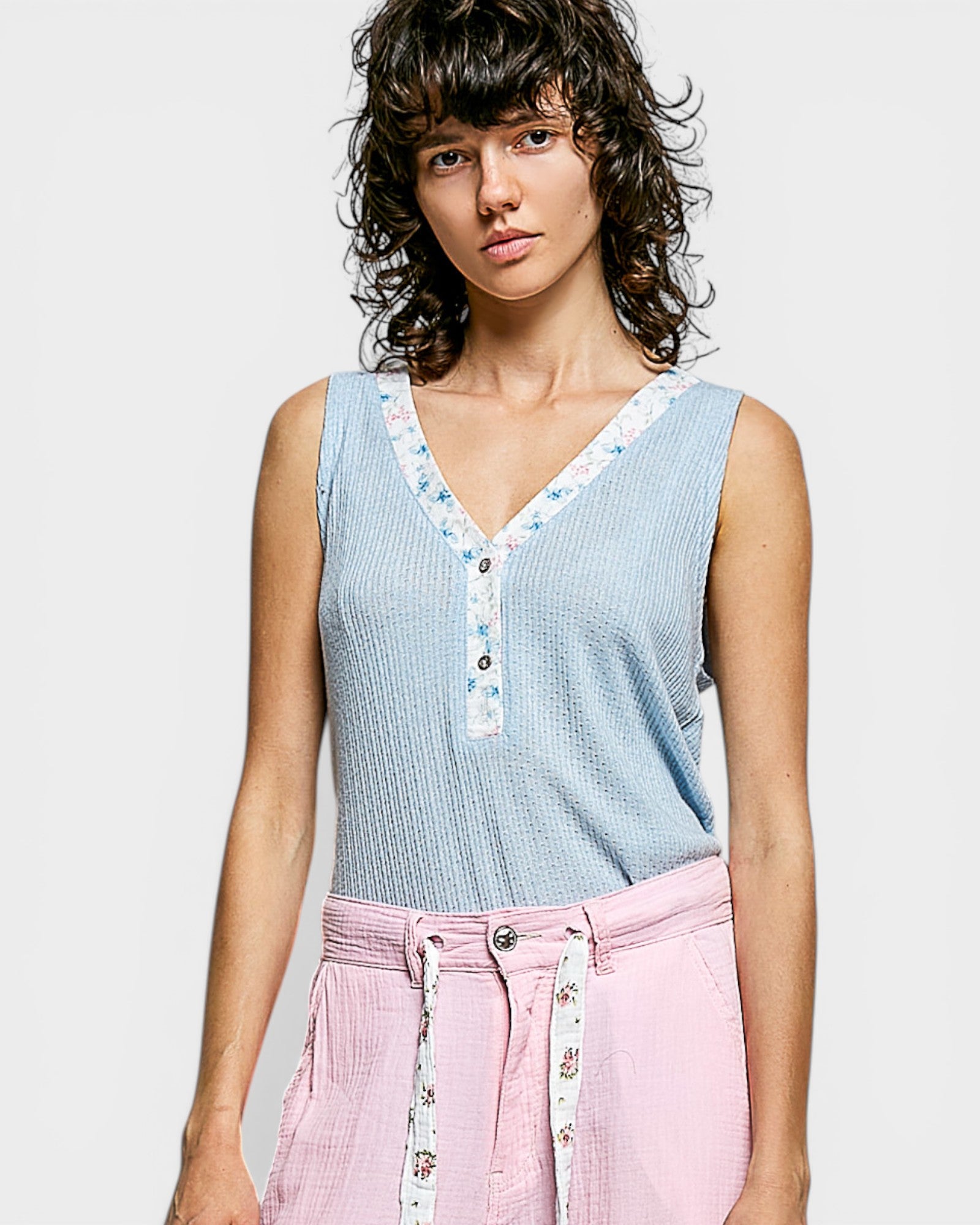 Woman wearing a light blue sleeveless top with floral details and pink pants on a plain background