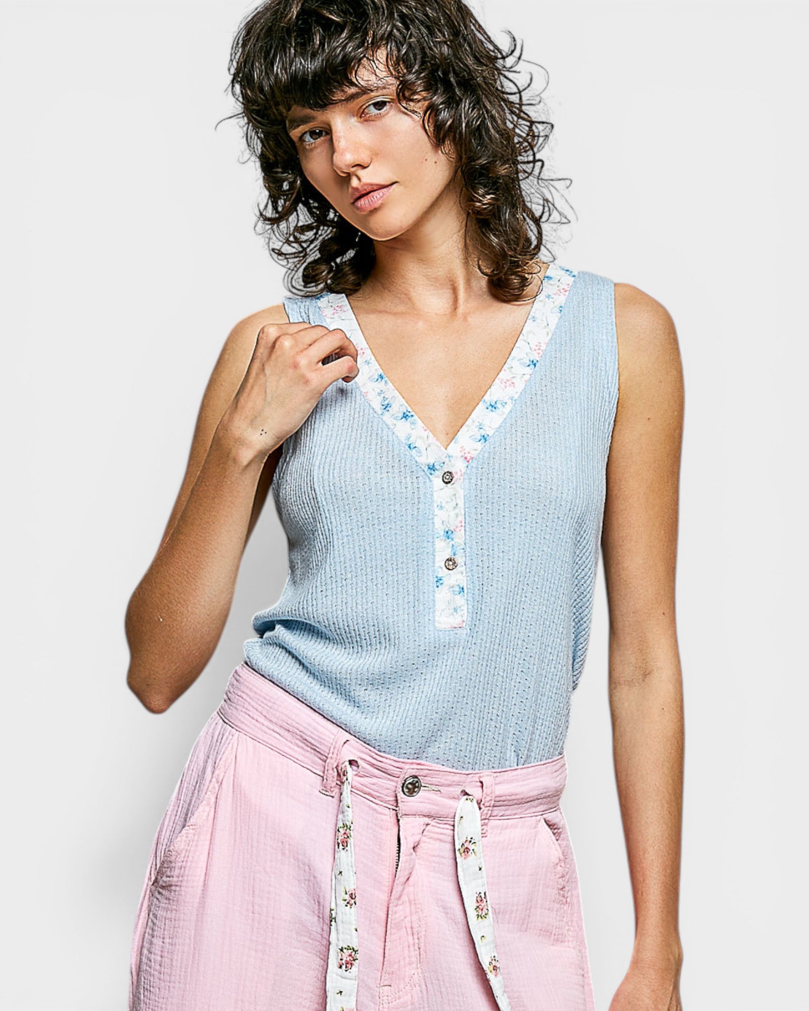 Woman wearing a light blue sleeveless top with floral trim and pink pants against a white background