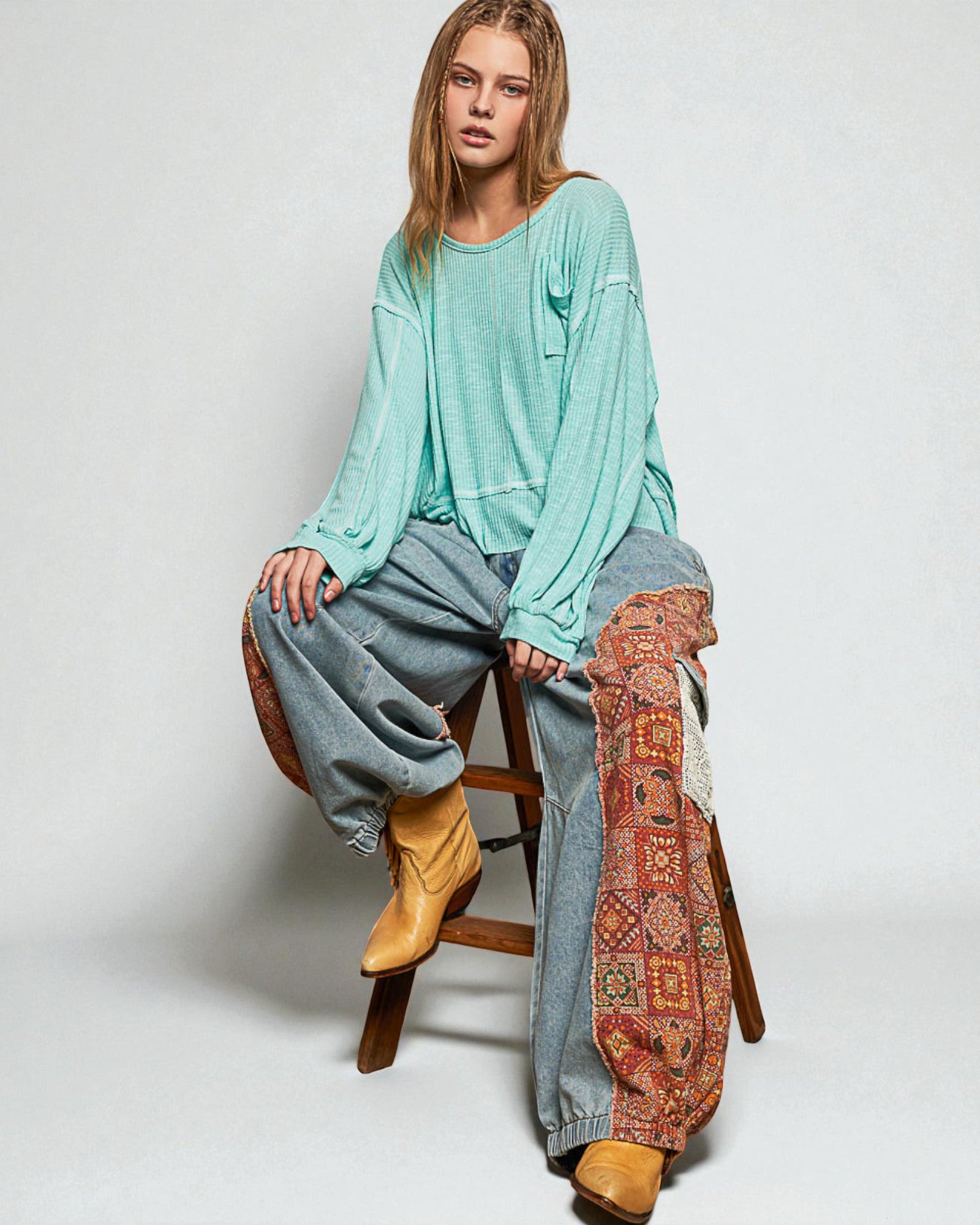 Woman sitting on a wooden stool wearing a teal sweater, gray pants, and yellow boots with a patterned blanket.