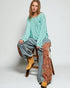 Woman sitting on a wooden stool wearing a teal sweater, gray pants, and yellow boots with a patterned blanket.