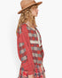 A woman in profile wearing a red and beige plaid patchwork cardigan with a cream lace tie and a matching plaid skirt, styled with a tan wide-brim hat against a plain white background.