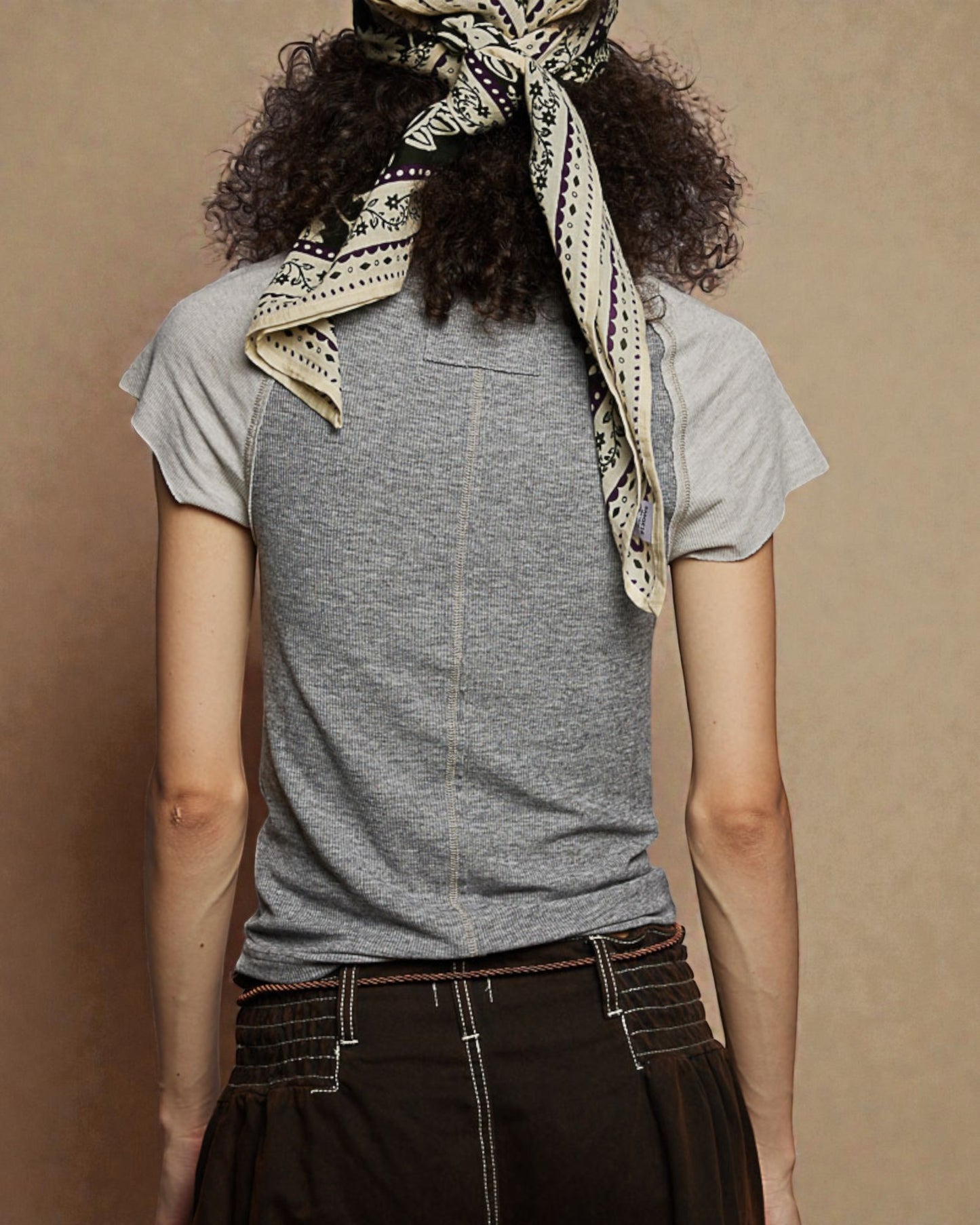 Person wearing a gray t-shirt and dark pants with a patterned scarf on a beige background