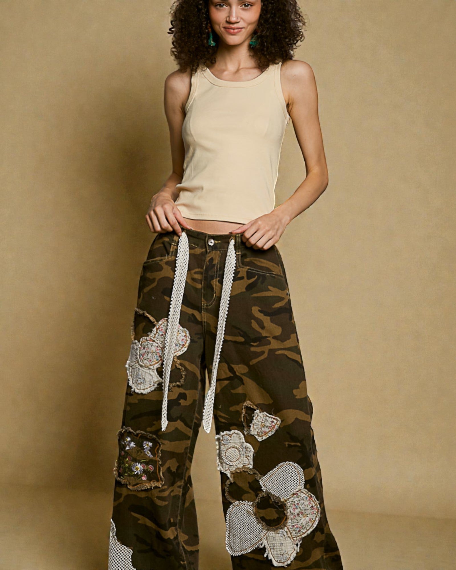 Woman wearing a beige tank top and camouflage pants with white lace details against a brown background