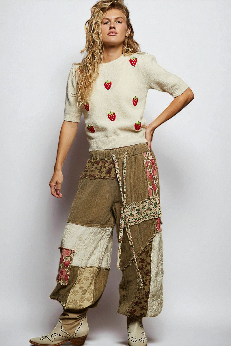 Woman wearing a cream sweater with red strawberry patterns and patchwork pants on a white background
