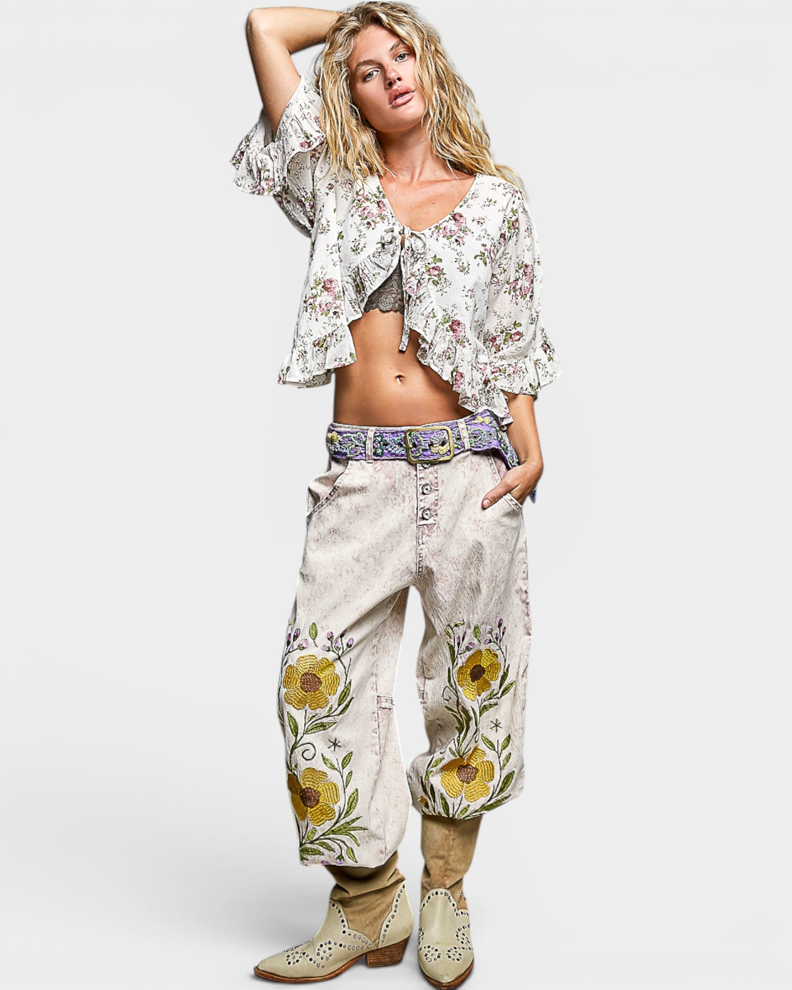 Woman wearing a floral top and embroidered pants with cowboy boots on a white background