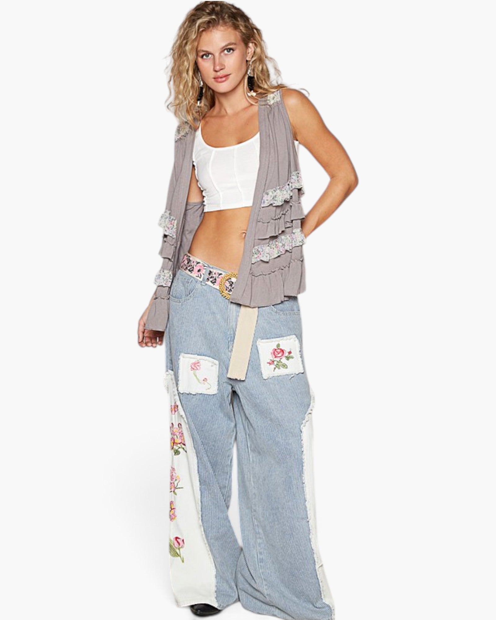 A woman modeling a bohemian-style outfit featuring light blue wide-leg jeans with floral embroidered patches, a white crop top, and a taupe ruffled cardigan vest, set against a plain white studio background.