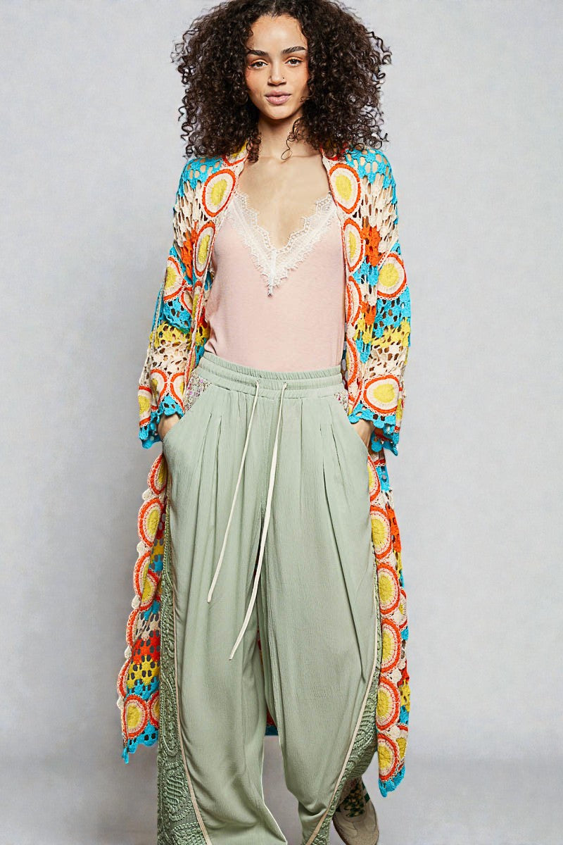 Woman wearing a colorful kimono-style jacket over a pink top and light green pants on a gray background