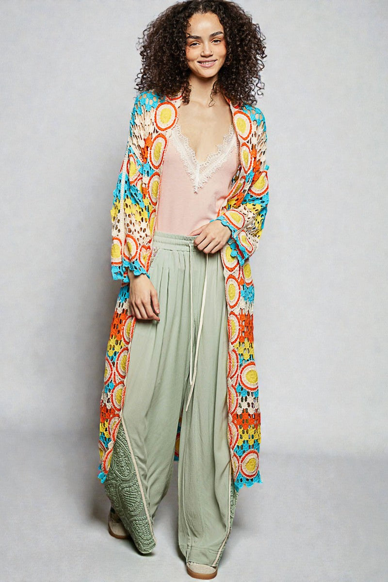 Woman wearing a colorful kimono-style cardigan over a light top and green pants on a plain background