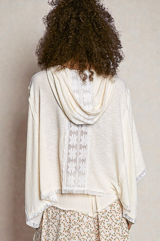 Person wearing a light-colored blouse with floral patterns and a scarf, standing against a plain background.