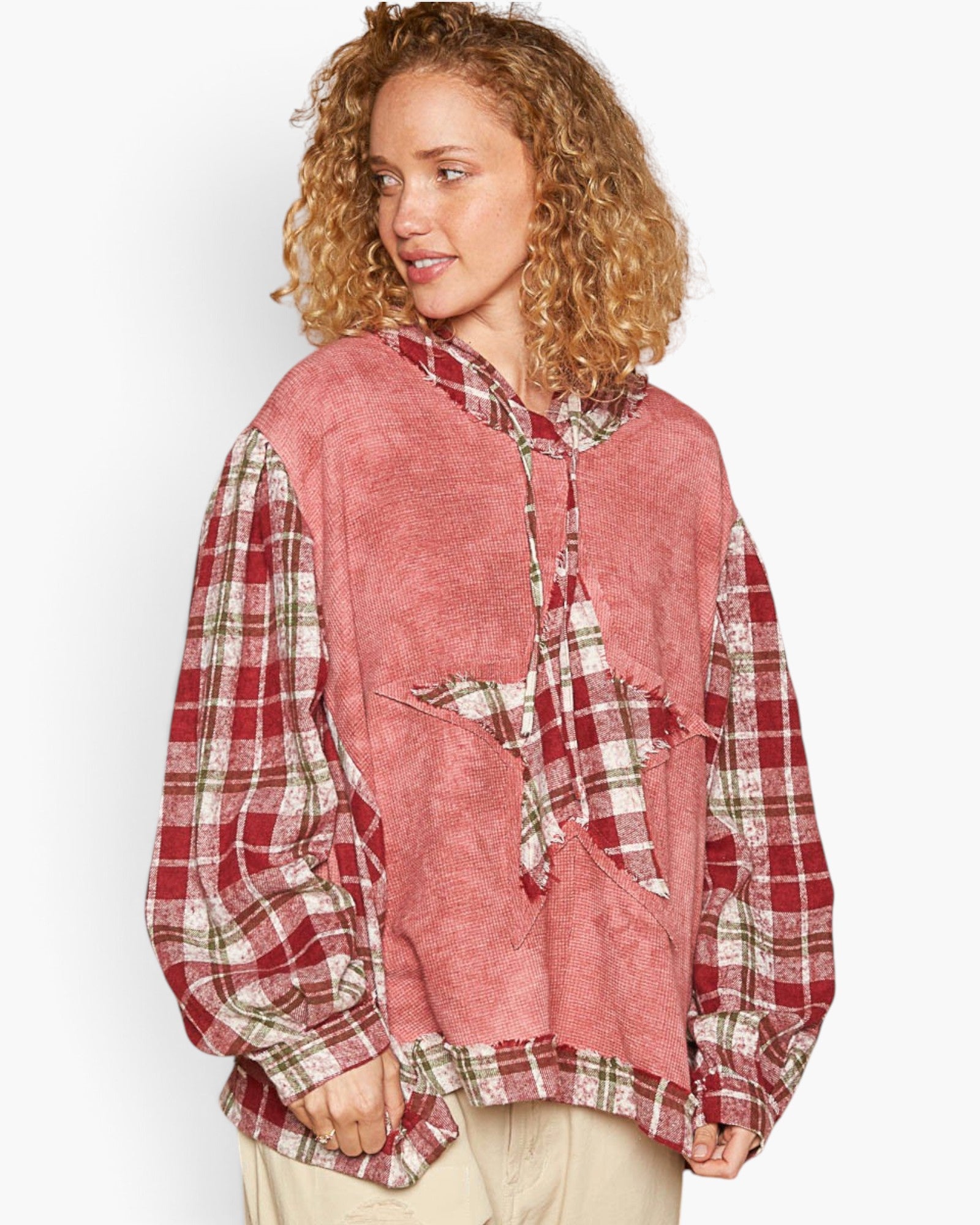 A woman wearing a unique patchwork hoodie with a dusty pink textured front panel and red, green, and white plaid sleeves and hood, paired with beige pants against a light gray background.