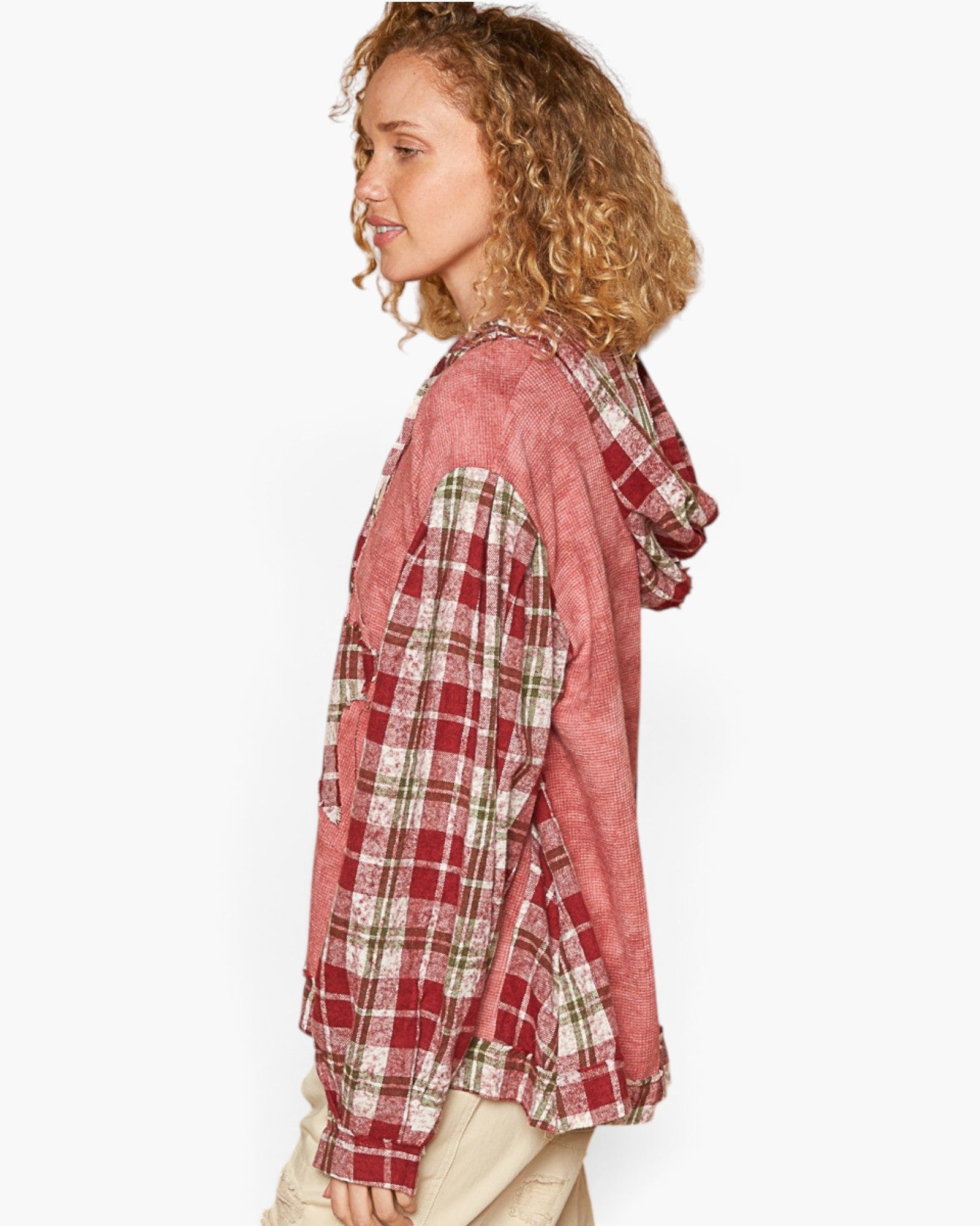 A woman shown in side profile wearing a hooded pullover top with a color-blocked design: a dusty rose hood and body, and red, cream, and green plaid sleeves. She is also wearing light beige pants against a clean white studio background.
