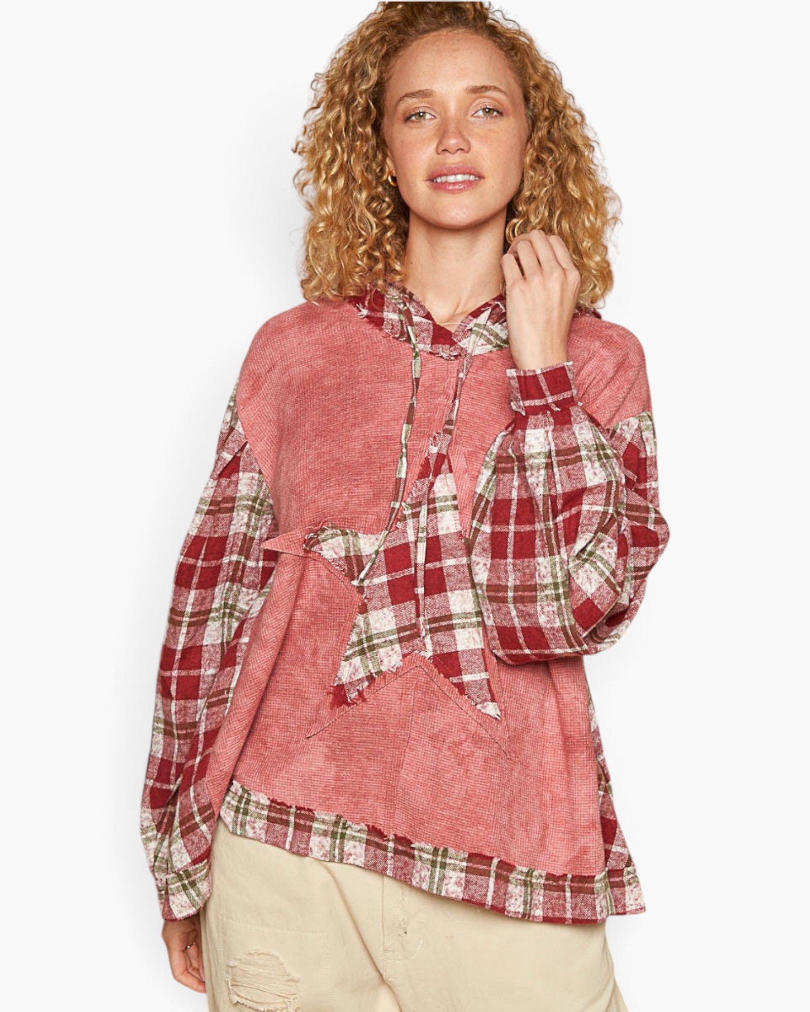 A woman modeling a long-sleeve hooded top with a dusty pink body and red, green, and white plaid panels on the sleeves, hood, and hem, paired with beige pants against a light gray studio background.