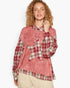 A woman modeling a long-sleeve hooded top with a dusty pink body and red, green, and white plaid panels on the sleeves, hood, and hem, paired with beige pants against a light gray studio background.