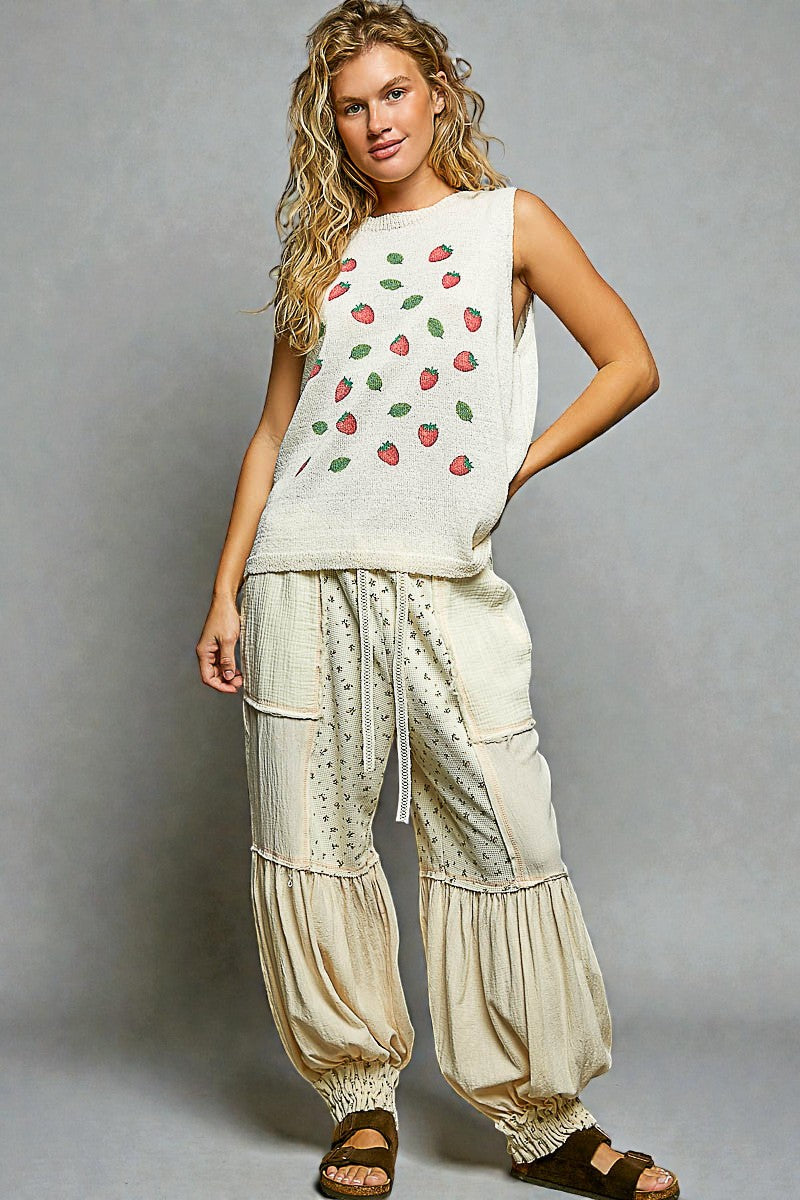Woman wearing a sleeveless top with strawberry pattern and loose beige pants on a gray background
