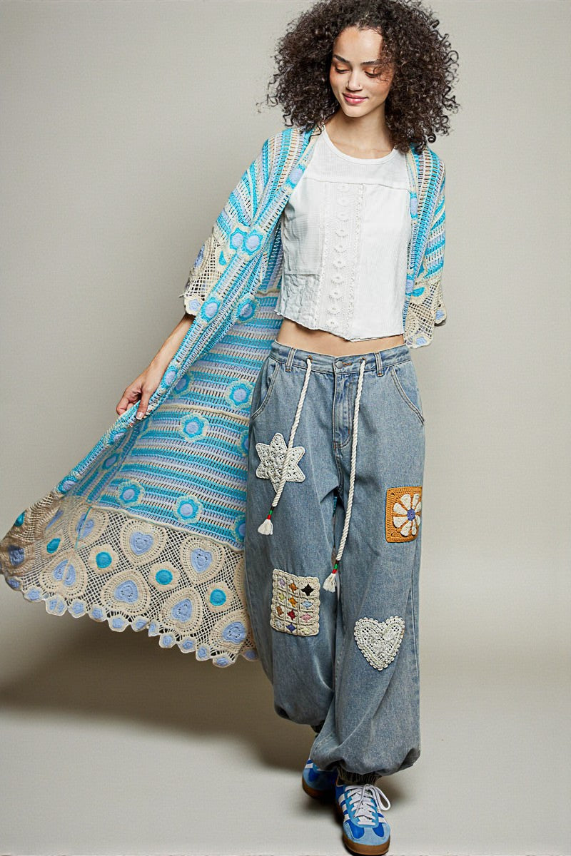 Woman wearing a blue embroidered shawl and denim pants with patches on a plain background